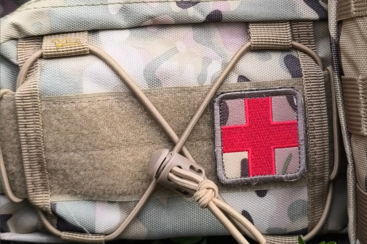 Close-up of a tactical pouch in a light camouflage pattern with tan webbing and a square patch featuring a red cross on a camouflage background.