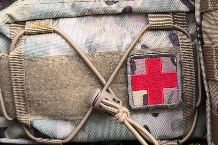 Close-up of a tactical pouch in a light camouflage pattern with tan webbing and a square patch featuring a red cross on a camouflage background.